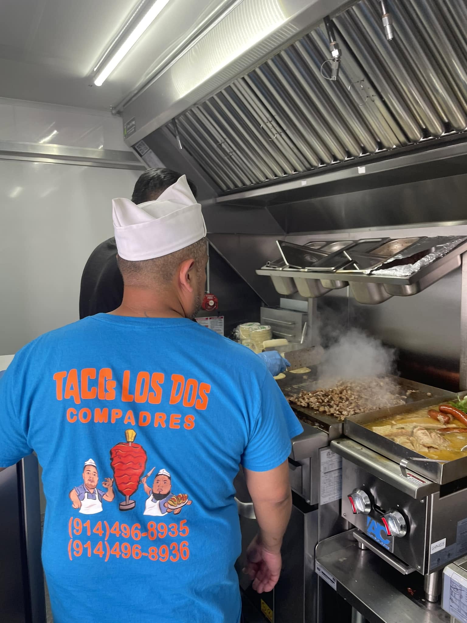 A cook in a Tacos Los Dos Compadres shirt grilling meat and warming tortillas on the truck's flat-top