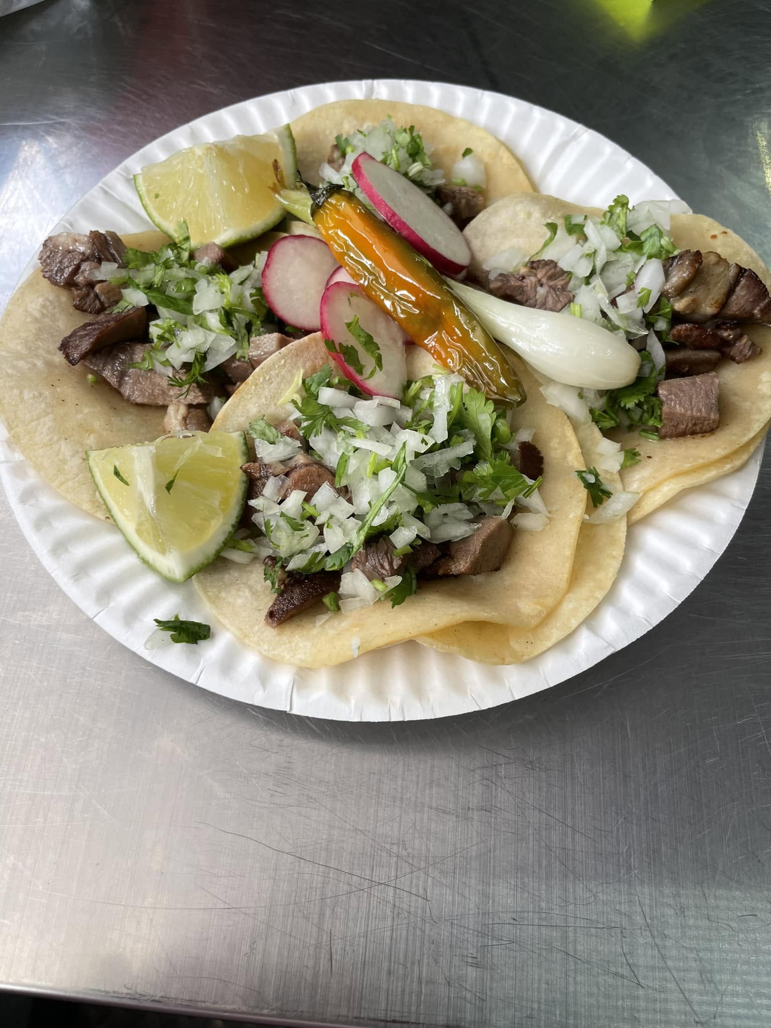 Steak tacos on a paper plate topped with cilantro, onion, radish, a grilled chile, and lime