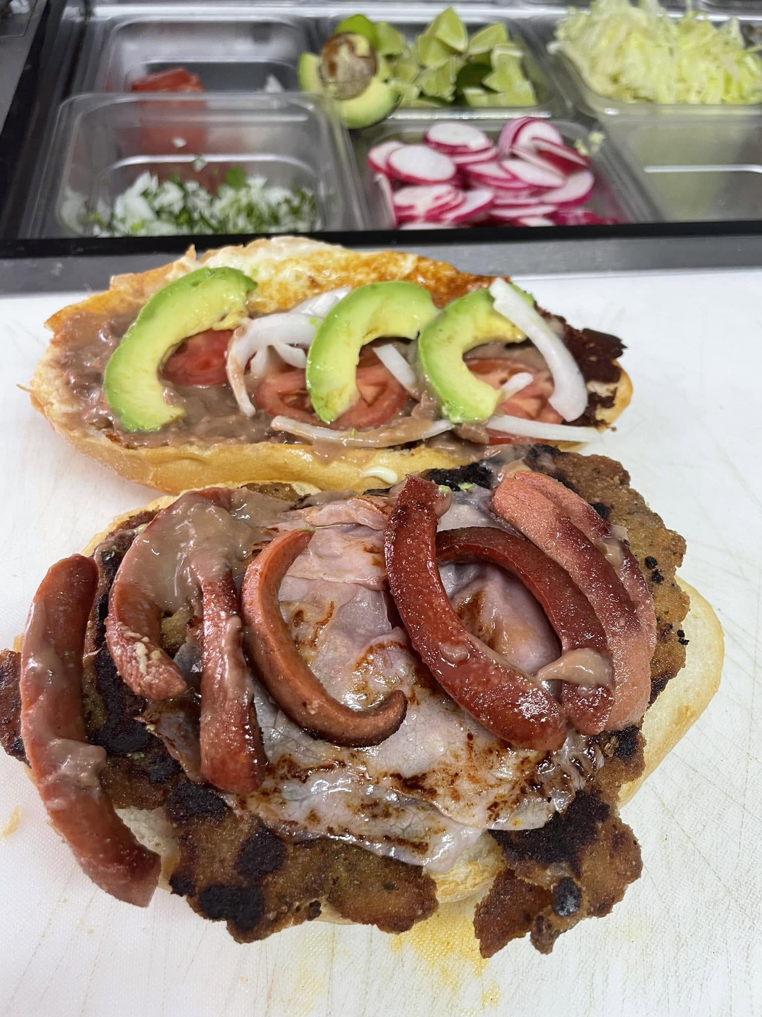 A loaded torta stacked with grilled meats beside a torta with refried beans, tomato, and avocado