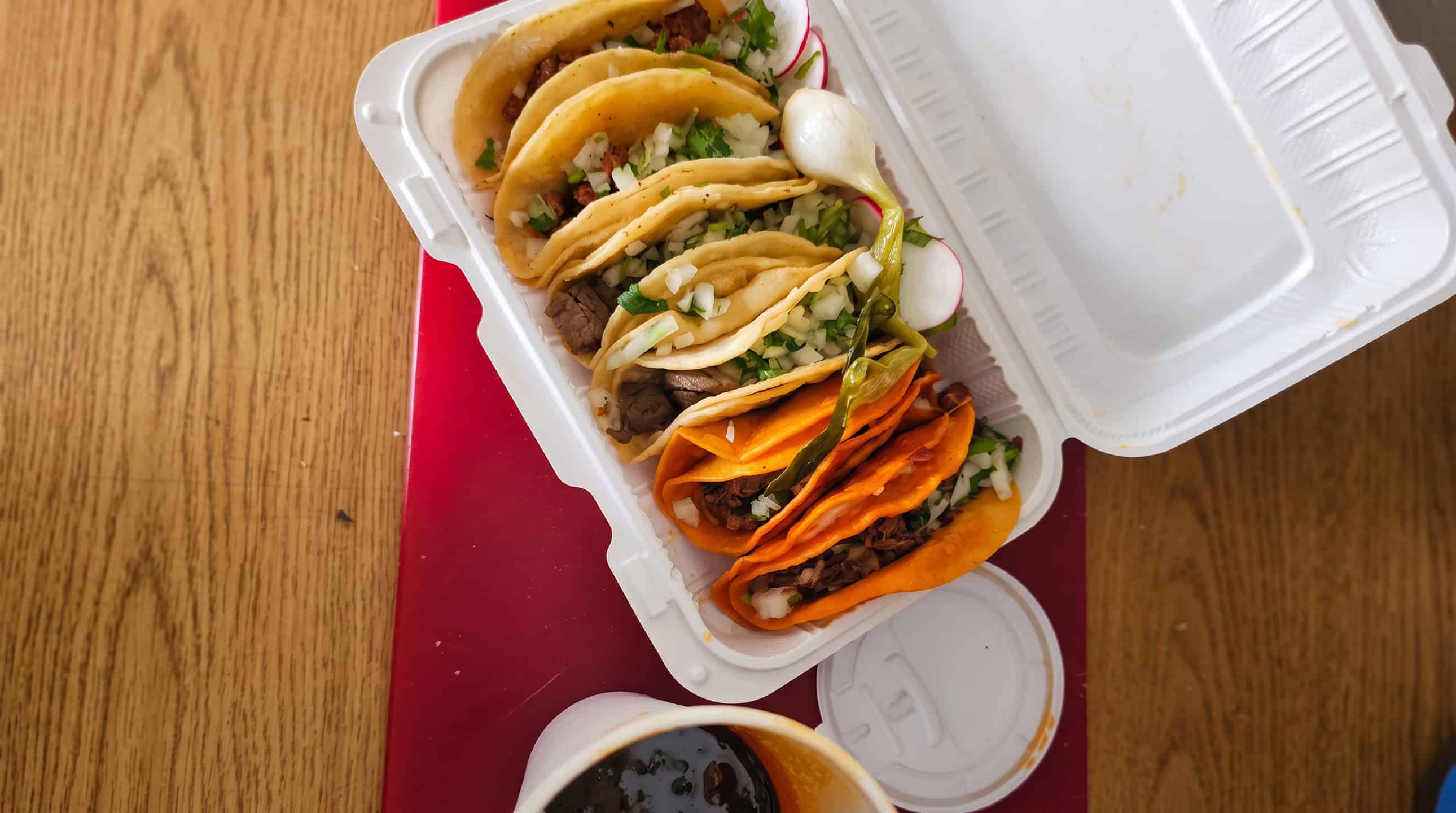 An overhead view of a takeout tray of steak, pastor, and birria tacos topped with onion and cilantro, served with radish, grilled onion, a roasted chile, and a side of salsa
